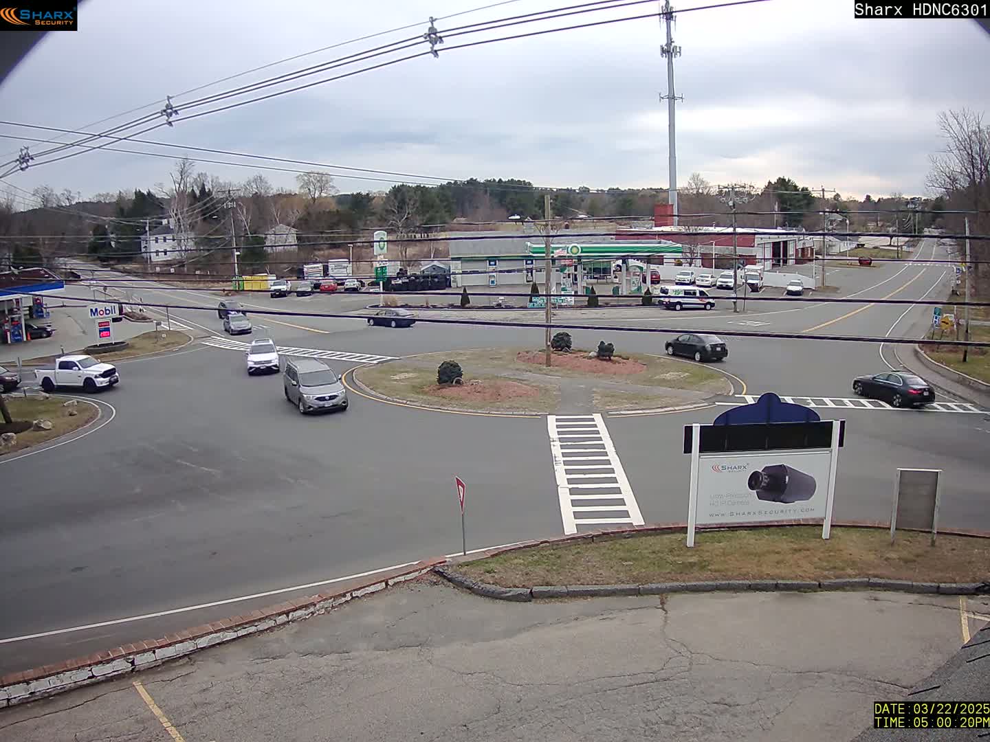 Several cars are driving around a traffic circle near a gas station on an overcast day.