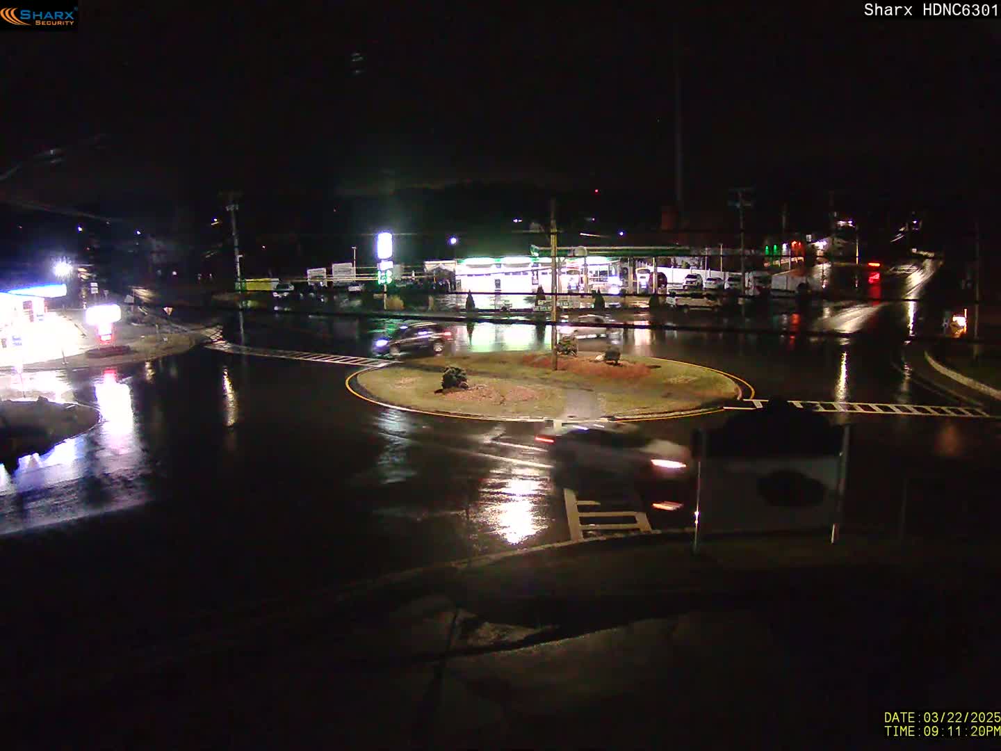 A rain-slicked roundabout at night, with a few cars visible and nearby buildings illuminated.