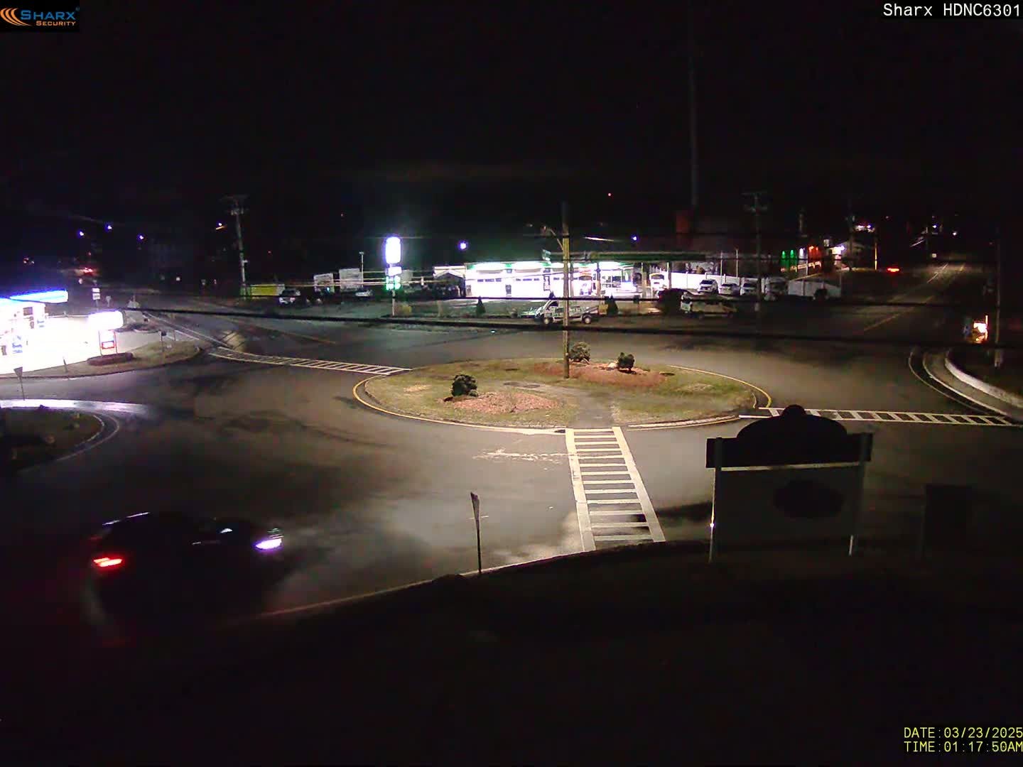 A dark-colored car drives through a roundabout at night, near a gas station and other buildings under artificial light.