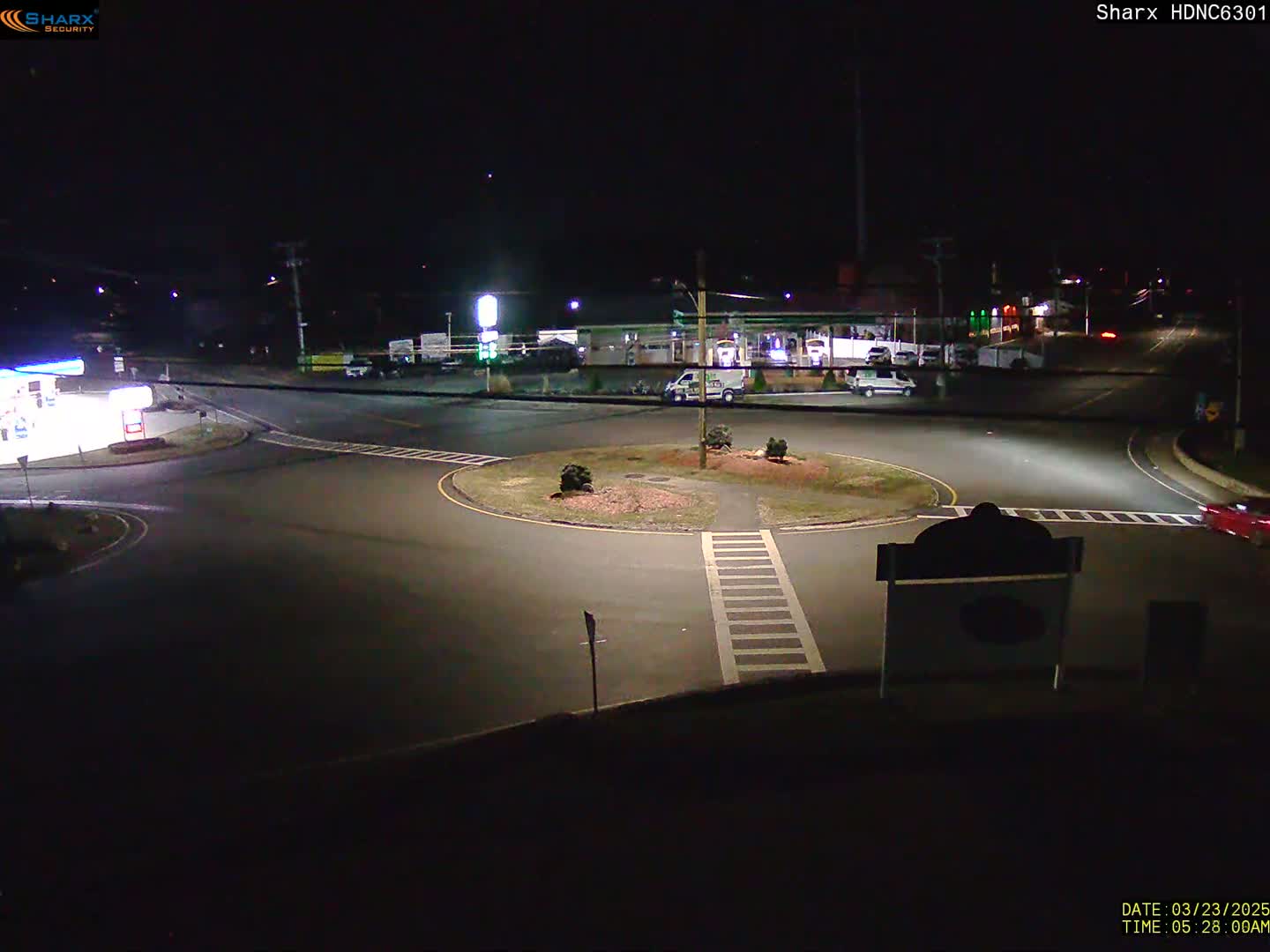 A nighttime view of a roundabout intersection near a gas station and other buildings, under clear skies.