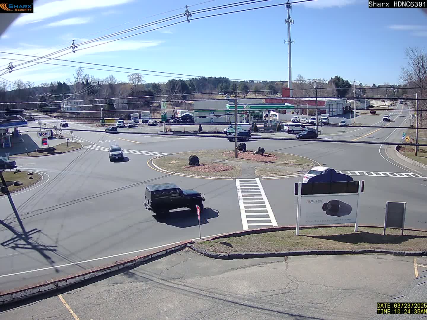 A sunny day shows a traffic circle with several vehicles, a gas station, and other businesses in the background.