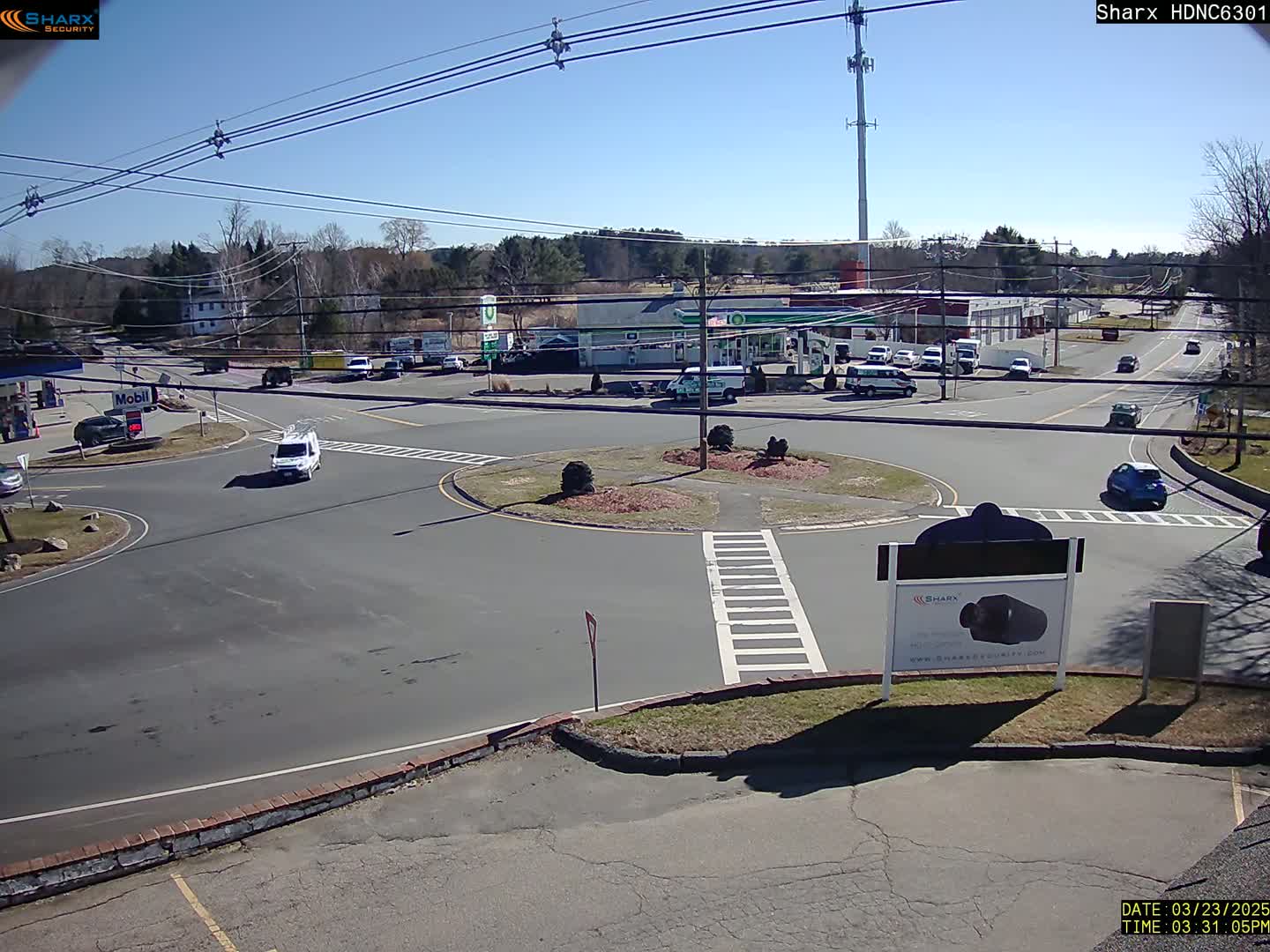 A sunny day shows a view of a roundabout intersection with several cars and buildings including a gas station, under a mostly clear sky.