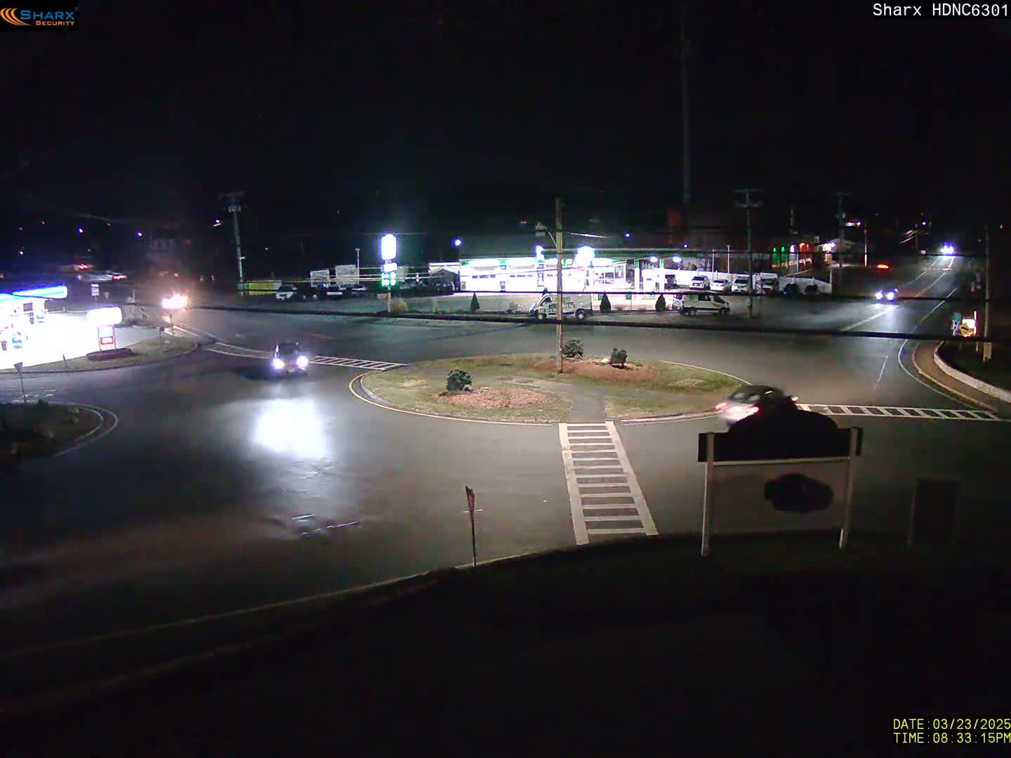 A nighttime view of a roundabout intersection with several vehicles and a gas station visible in the background under dark, clear skies.