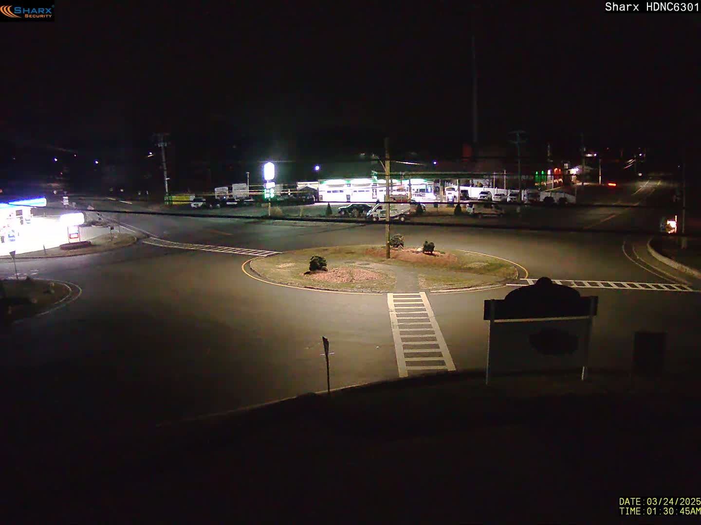 A nighttime view of a roundabout intersection near a gas station and other businesses under dark, clear skies.