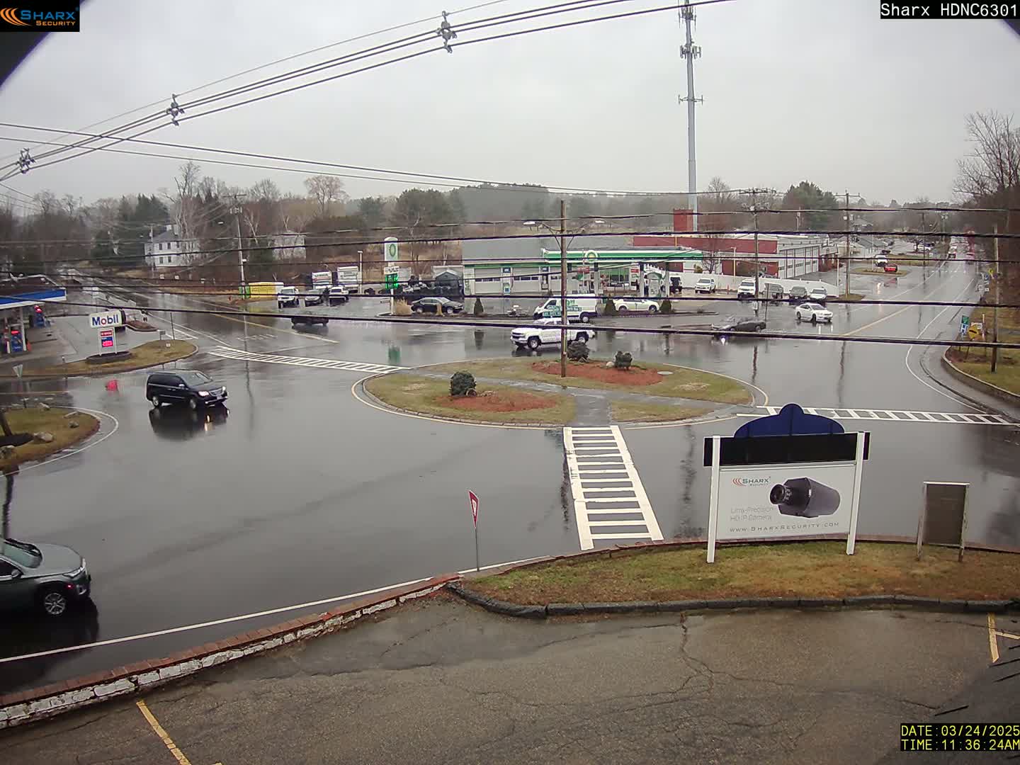 A wet, rainy scene shows a traffic circle with several cars and buildings including a gas station in the background.
