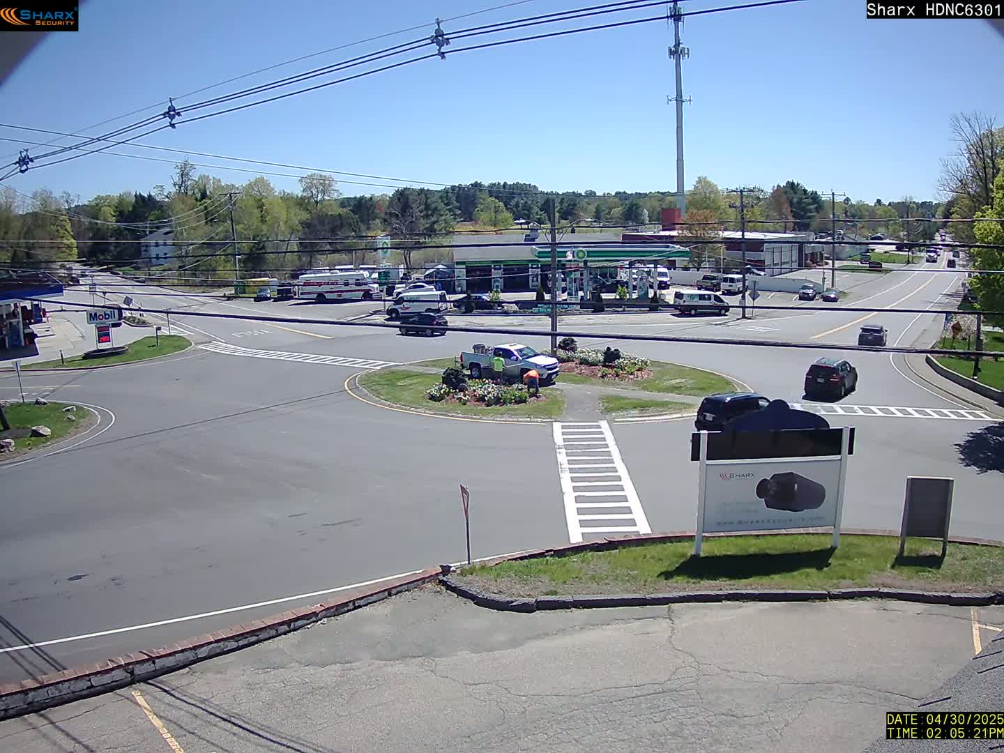 A sunny day shows a roundabout intersection with several vehicles, a gas station, and surrounding businesses.