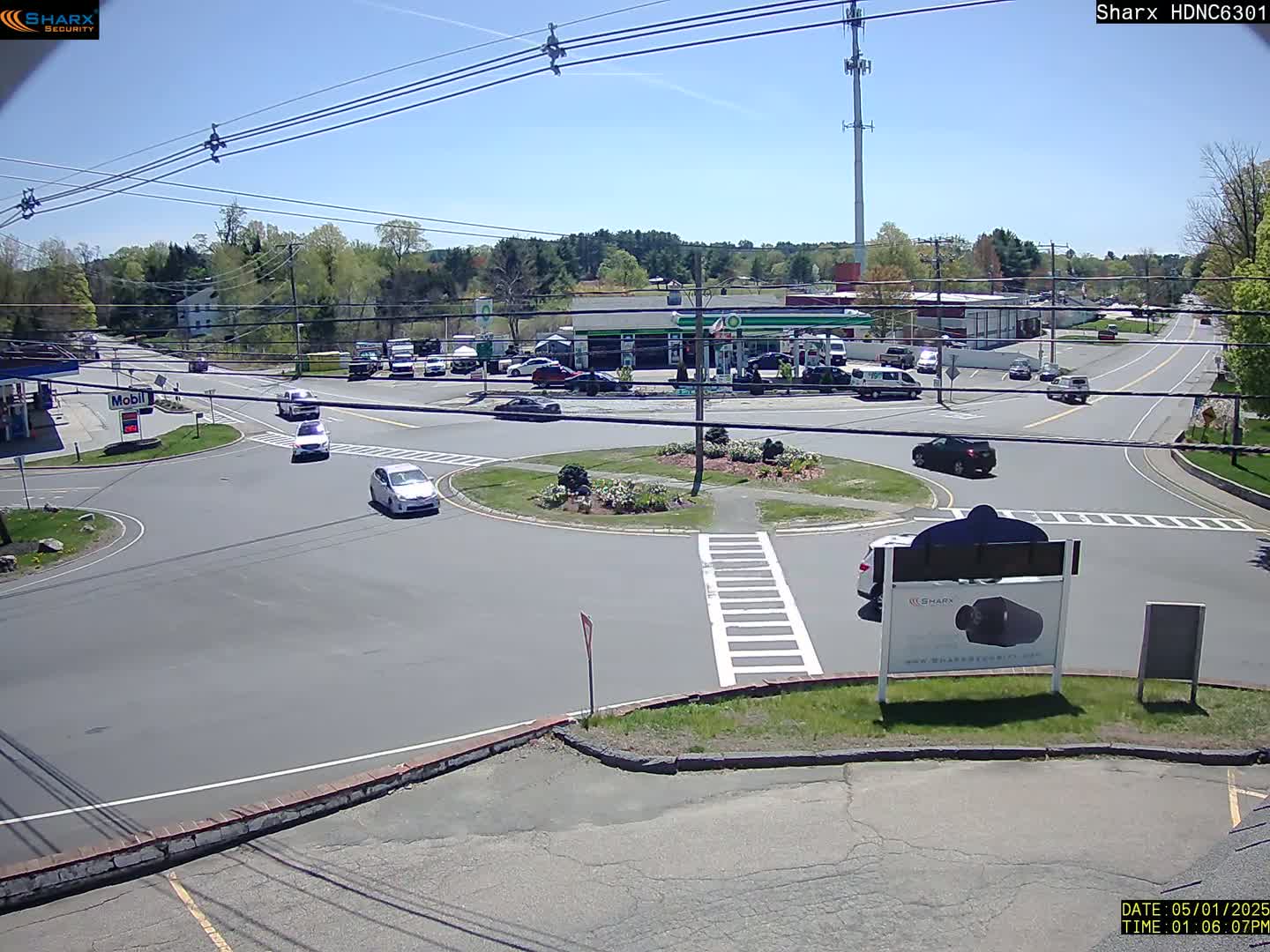 A sunny day reveals a traffic circle with several cars and a gas station in the background.
