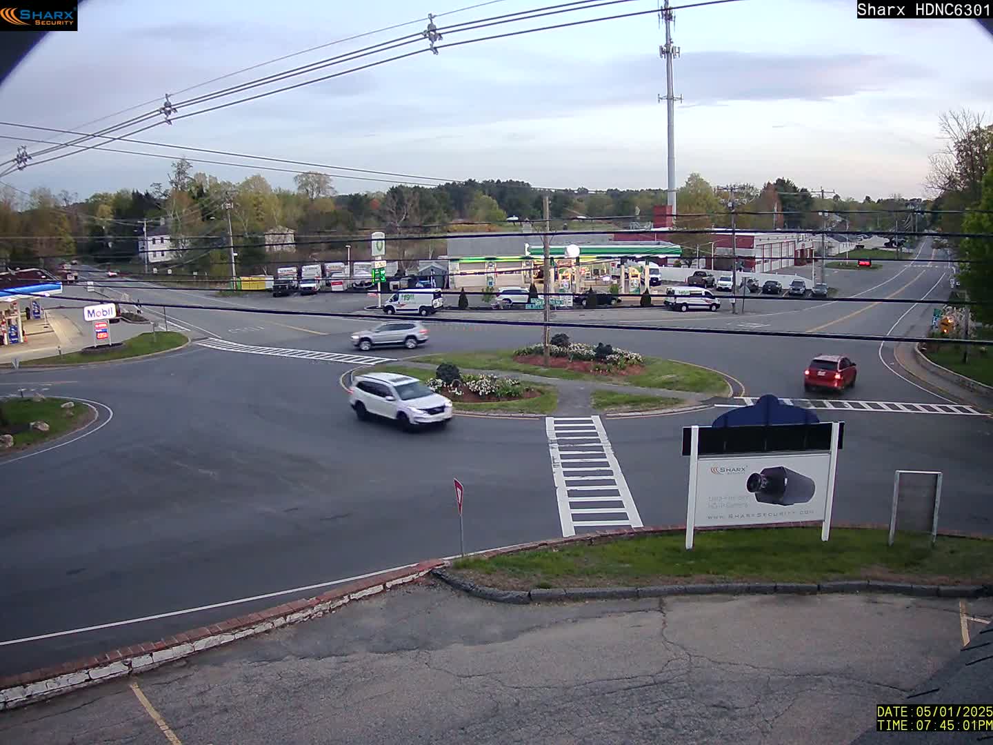 A mostly empty roundabout and intersecting roads are seen under a twilight sky, with several cars visible.