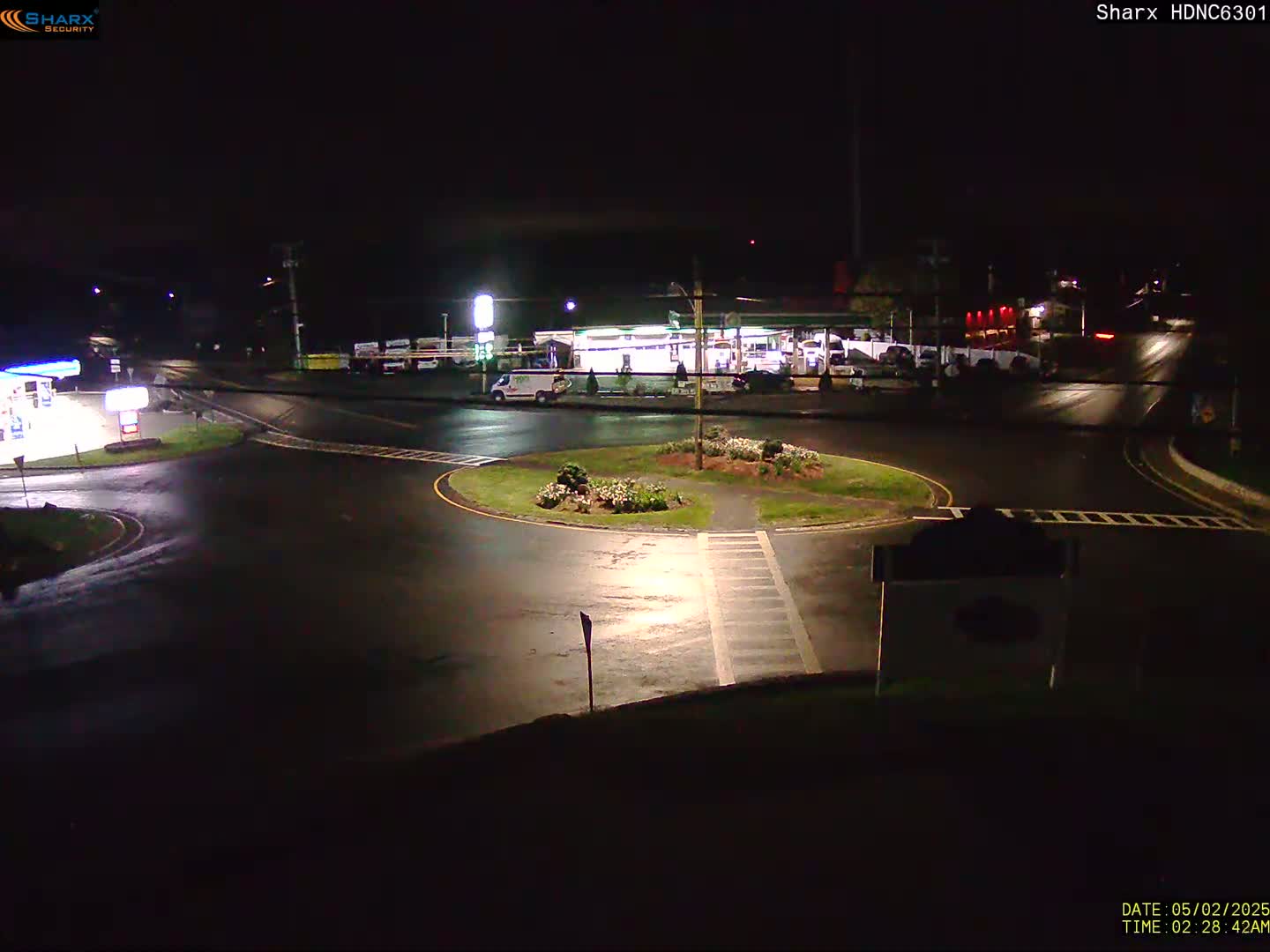 A wet, dark nighttime scene shows a circular intersection with a gas station and several vehicles visible, surrounded by minimal landscaping.