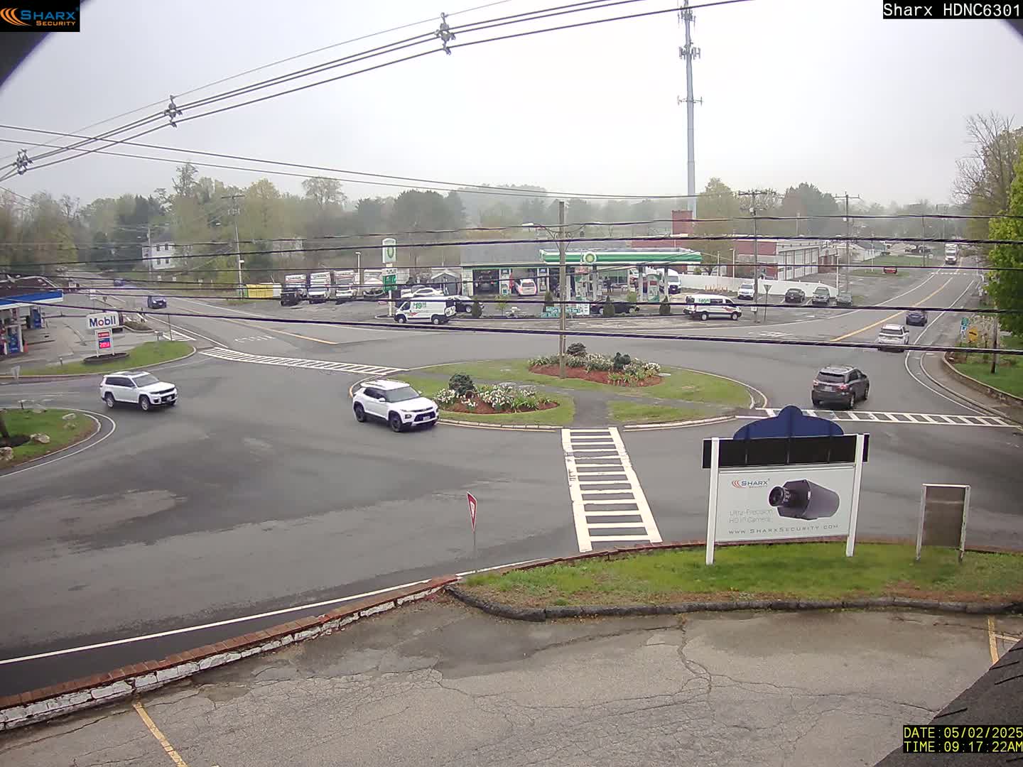 A slightly overcast view of a gas station and roundabout with several cars, surrounded by trees and buildings.