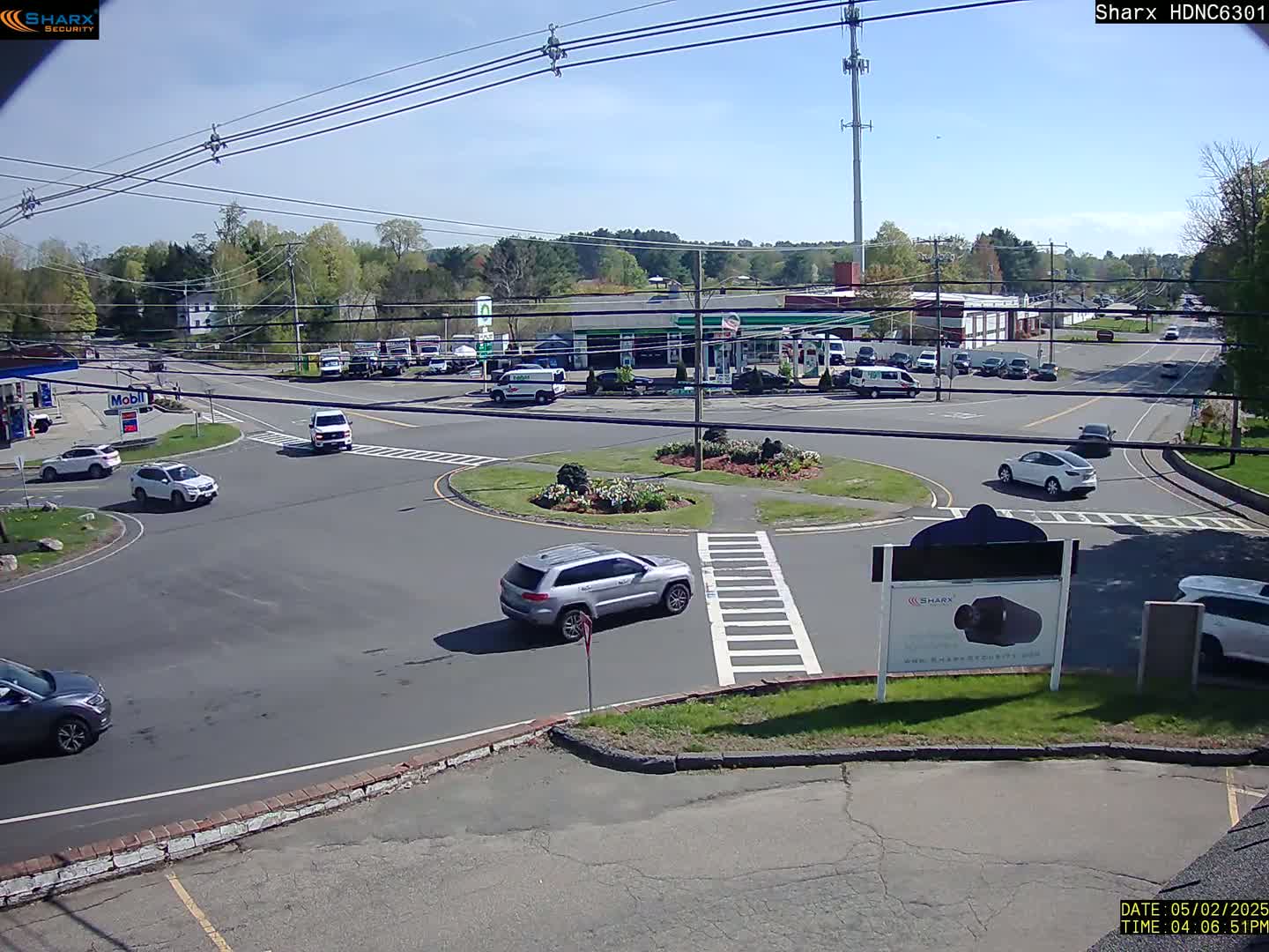 A sunny day reveals a traffic circle with several cars, surrounded by a gas station, businesses, and trees.