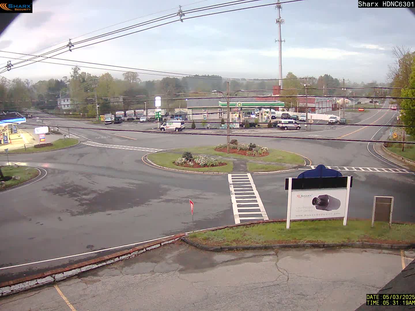 A wet roundabout in a small town, with a gas station and other buildings visible, under an overcast sky.