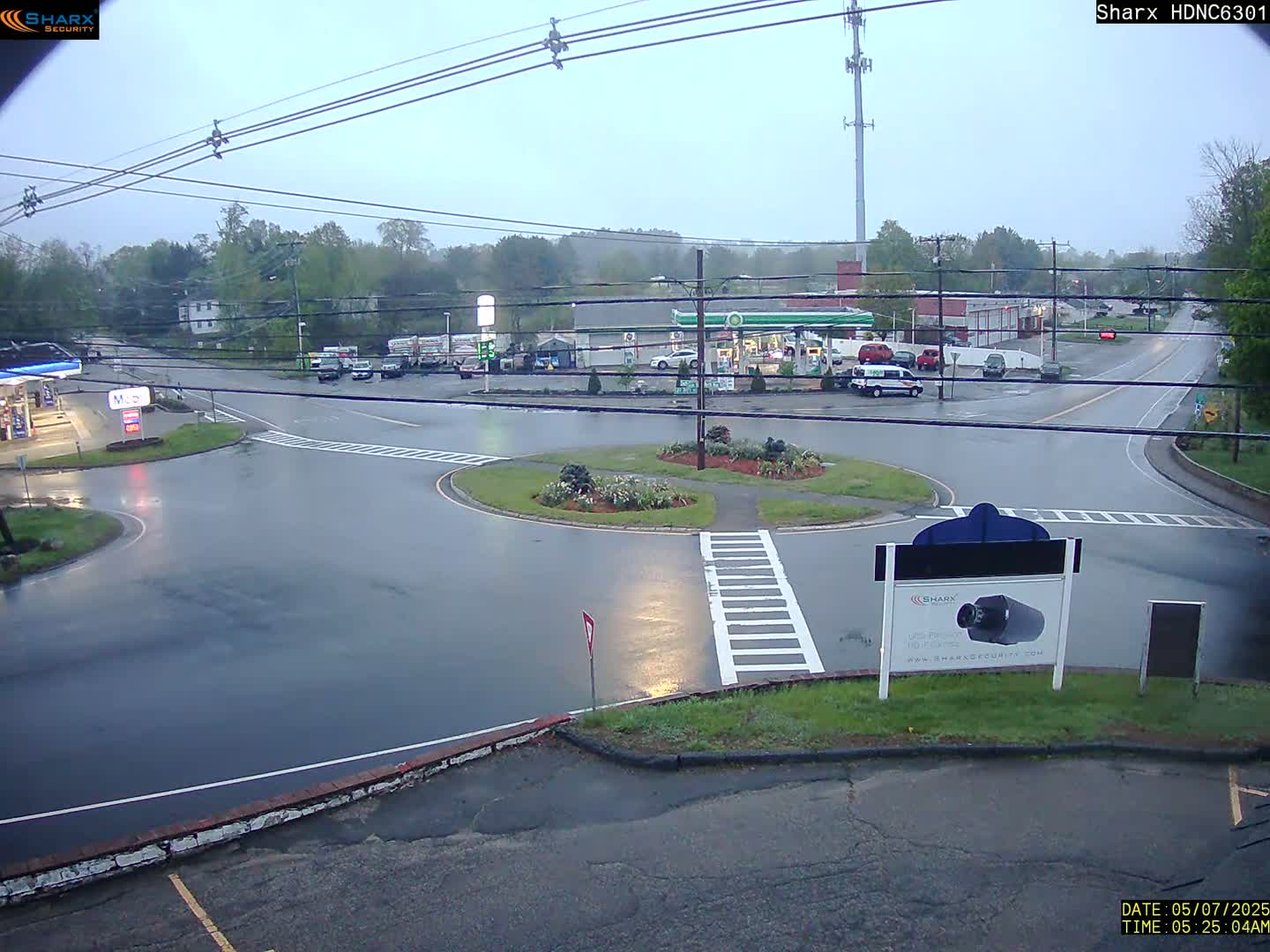 A wet roadway intersection with a gas station and several vehicles is seen on an overcast day.