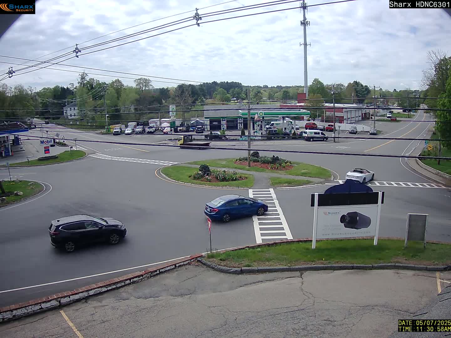 A mostly sunny day shows a gas station and several cars navigating a roundabout at a road intersection, surrounded by trees and other buildings.