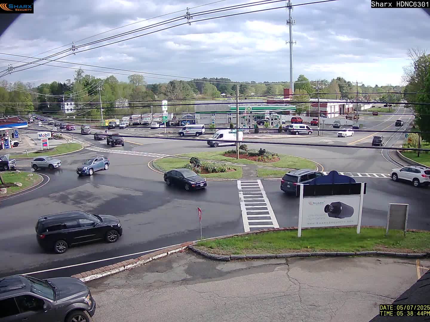 Several vehicles are navigating a roundabout and nearby roads under a partly cloudy sky.