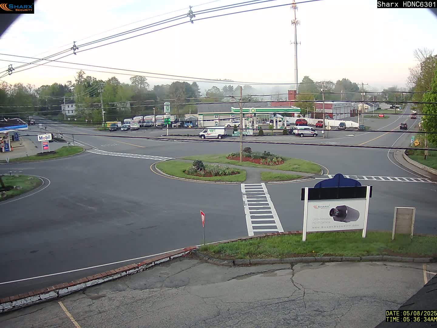A mostly clear, hazy morning view of a road intersection with a gas station and several vehicles, centered around a small roundabout.