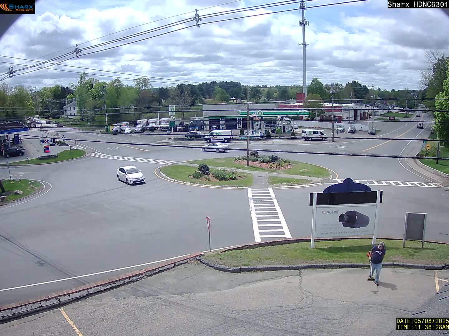 A partly cloudy day shows a roundabout intersection with several cars and trucks, a gas station, and other buildings, with a person standing near a sign in the foreground.
