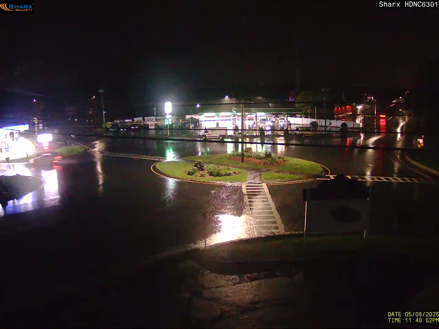 A rain-soaked roadway curves around a small, grassy island at night, with gas station lights reflecting in the puddles.