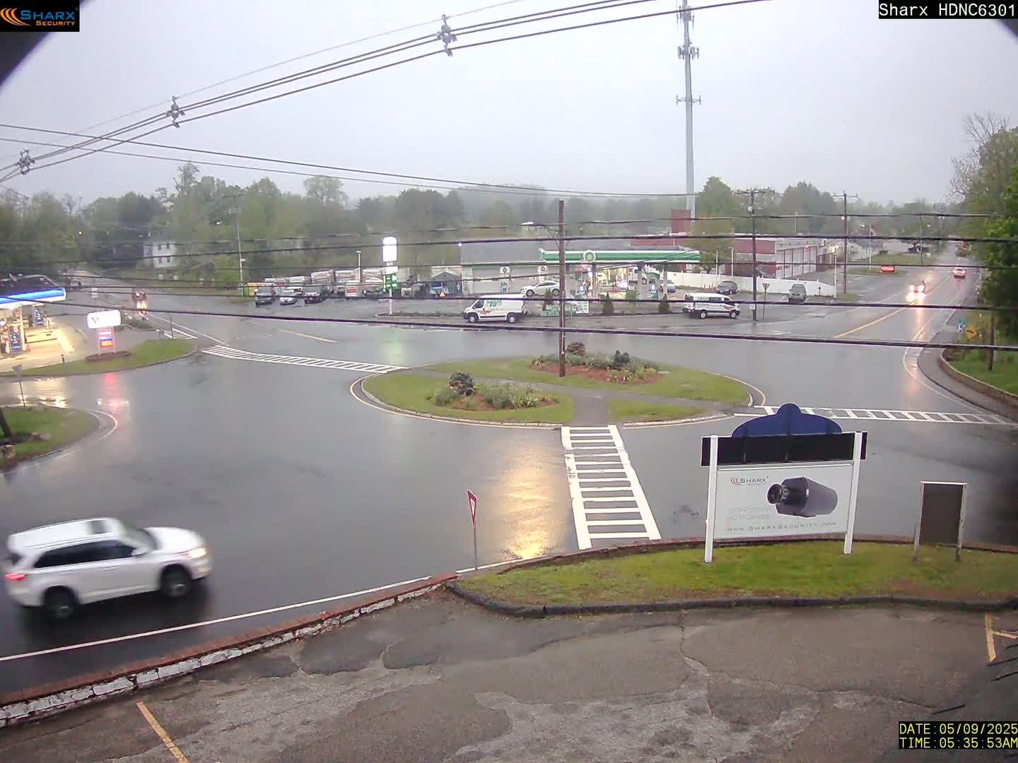 A wet roundabout with a few cars and gas station in the background during a light rain.