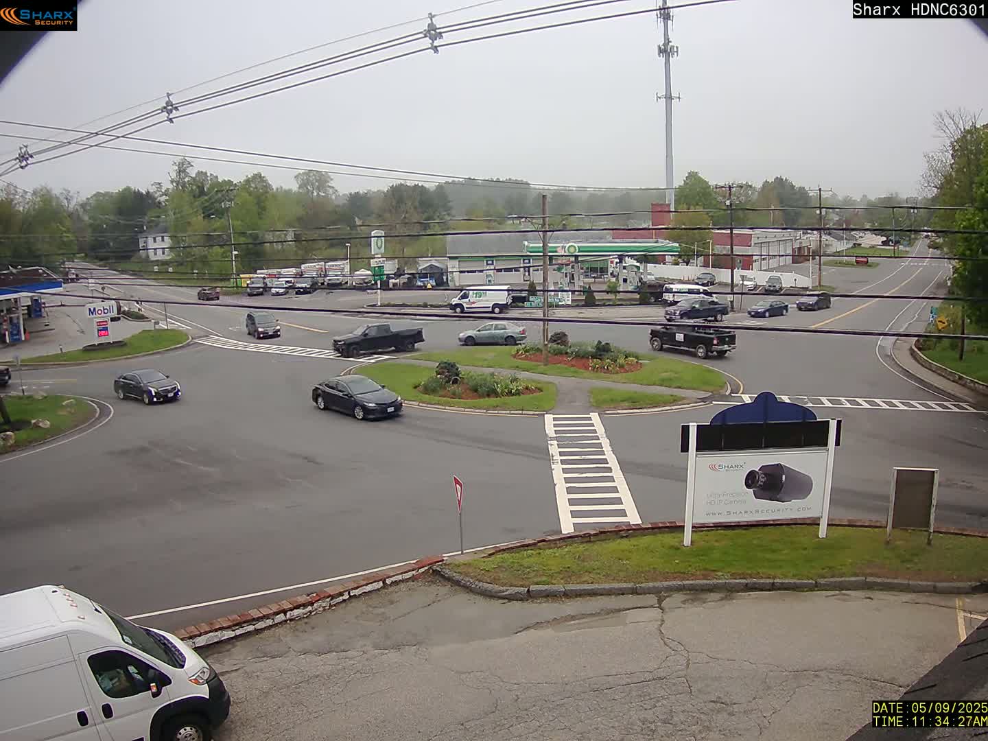 Several cars are driving around a roundabout in front of a gas station and other businesses on an overcast day.