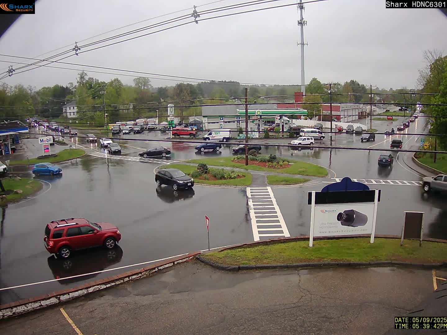 A wet, overcast day shows several cars navigating a roundabout and nearby roads, with gas stations and other buildings visible in the background.