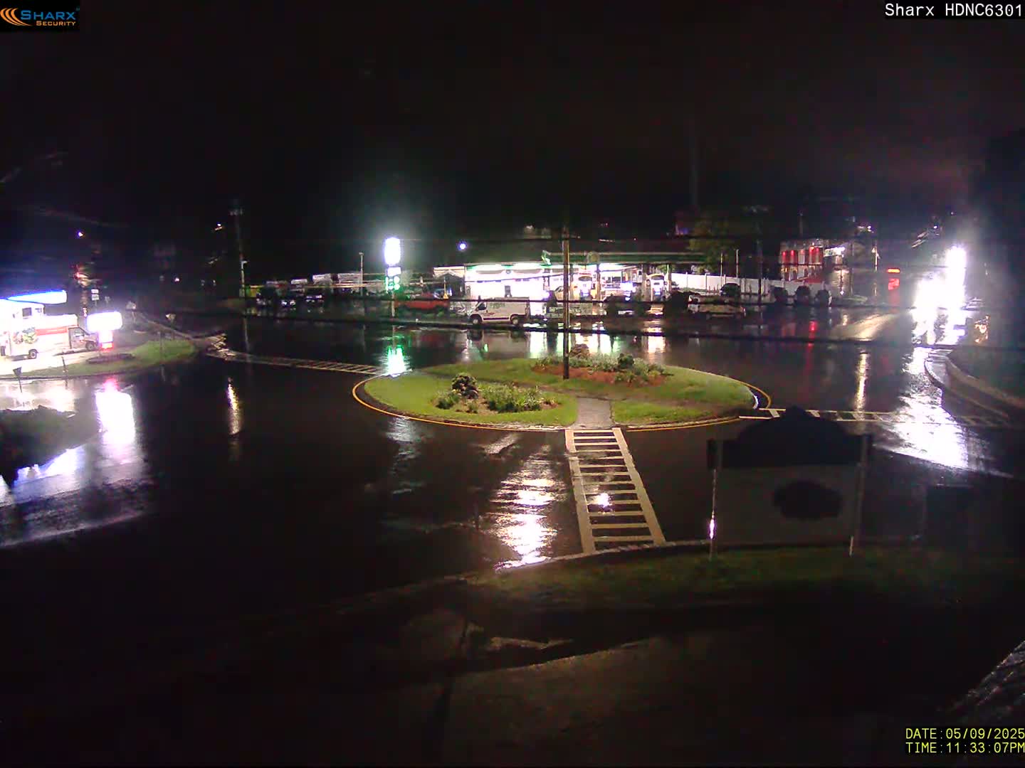 A rain-soaked roundabout at night, surrounded by buildings and a gas station, with reflections of lights in the wet pavement.