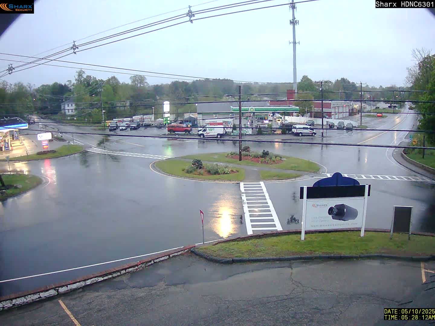 A wet, overcast scene shows a roundabout intersection with several vehicles parked near a gas station and a few cars traveling on the roads.