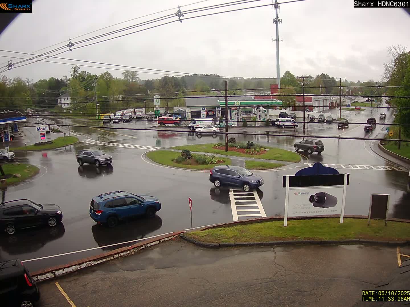 A rain-slicked roundabout with several cars and gas stations is visible in a parking lot near a wooded area.