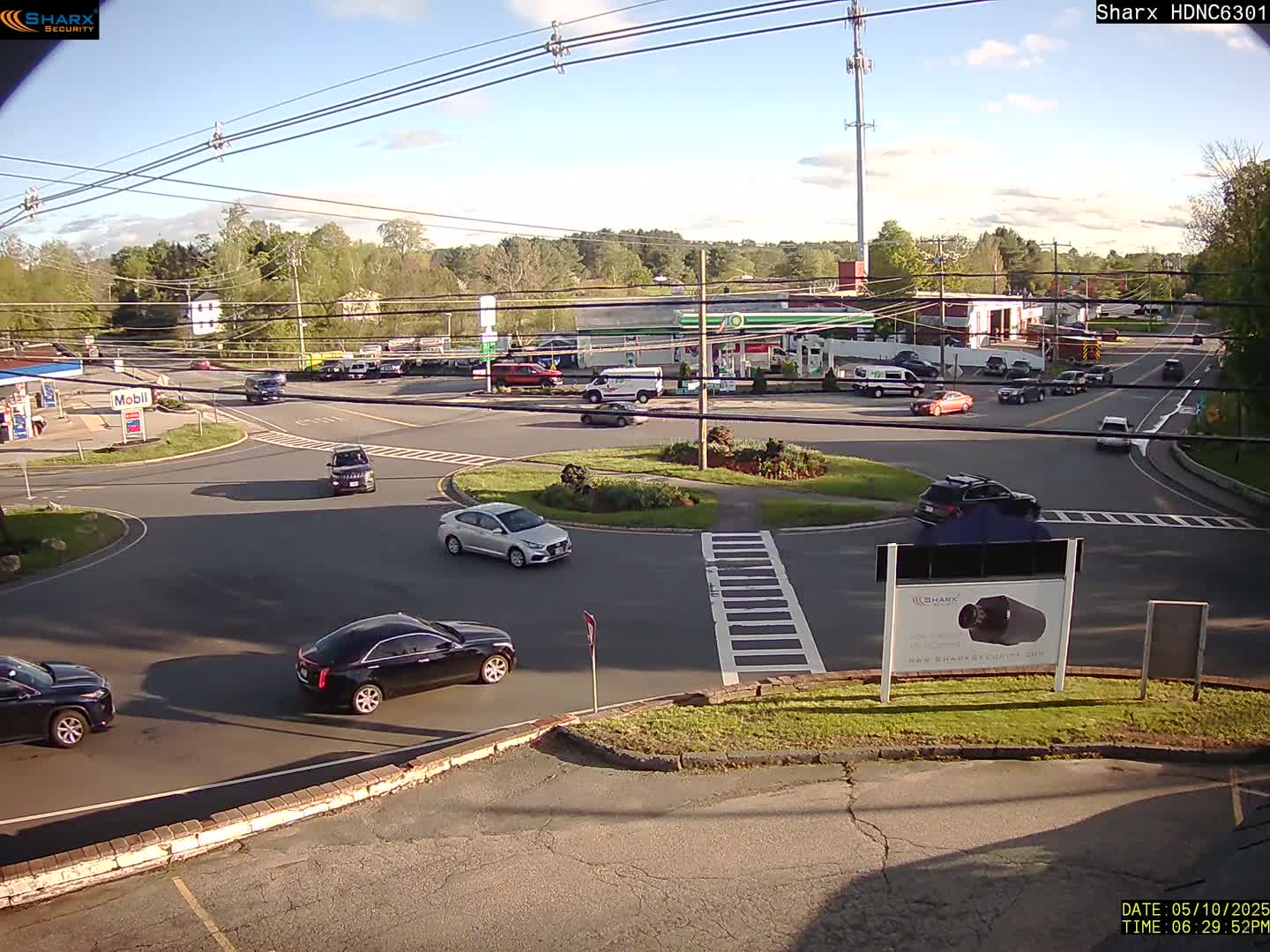 A sunny day at a gas station intersection shows several cars driving through a roundabout with other cars parked near nearby businesses.