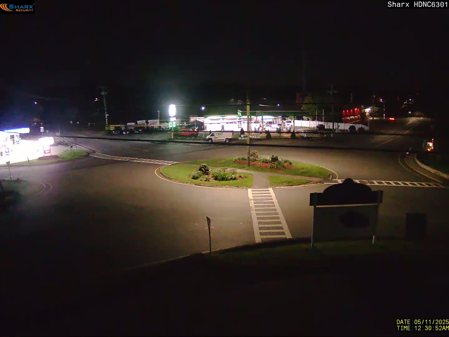 A nighttime view of a circular intersection with a gas station and other buildings in the background under clear skies.