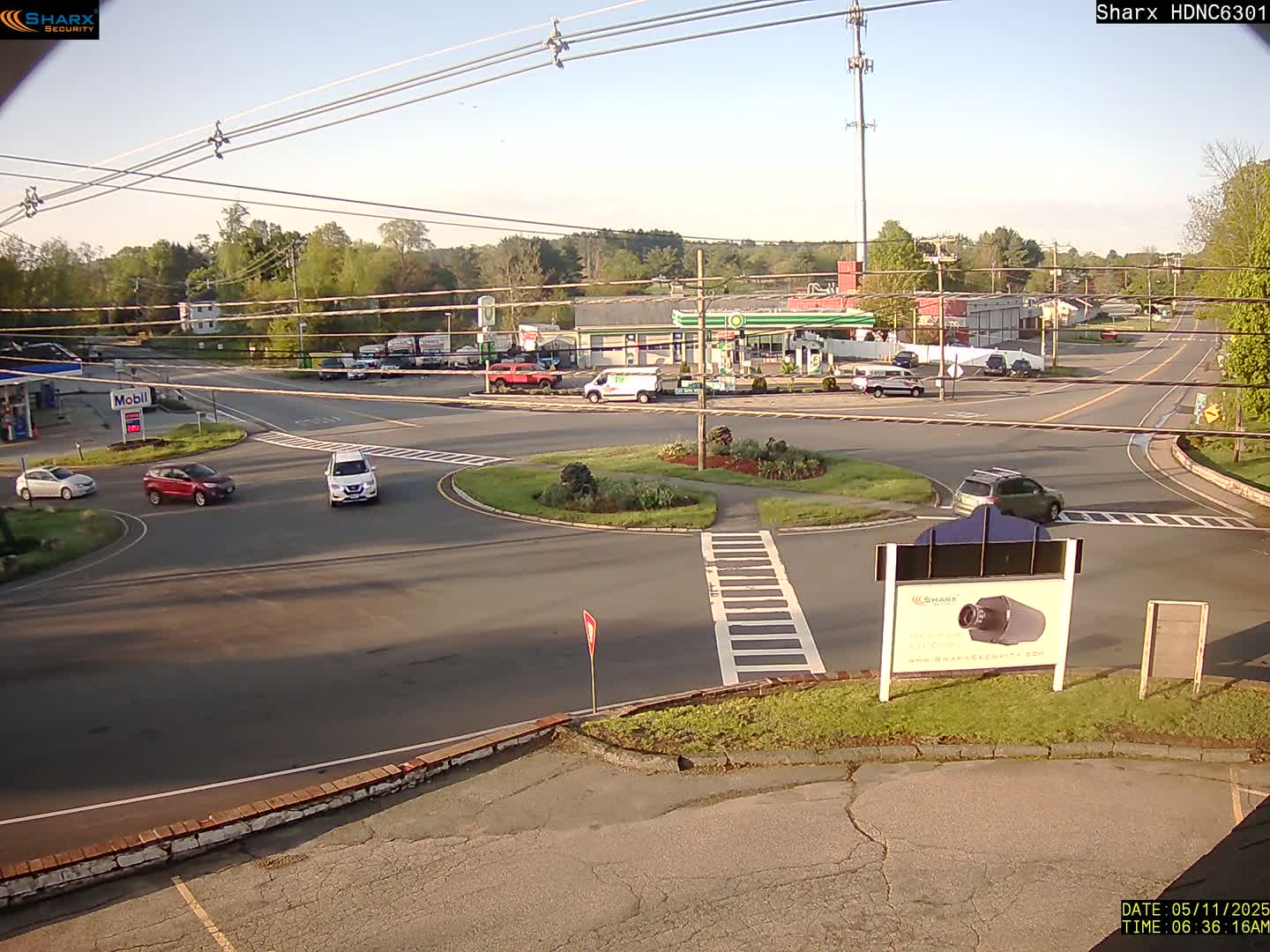 A sunny day reveals a mostly empty roundabout surrounded by a gas station, buildings, and several cars at an intersection.