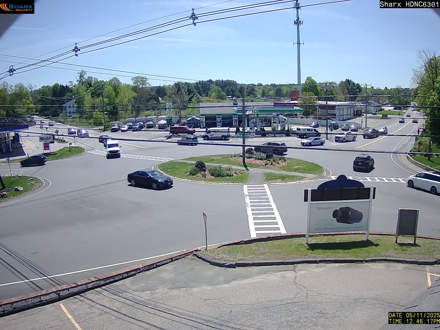 A sunny day shows a bird's-eye view of a roundabout intersection with several cars, a gas station, and surrounding buildings and trees.