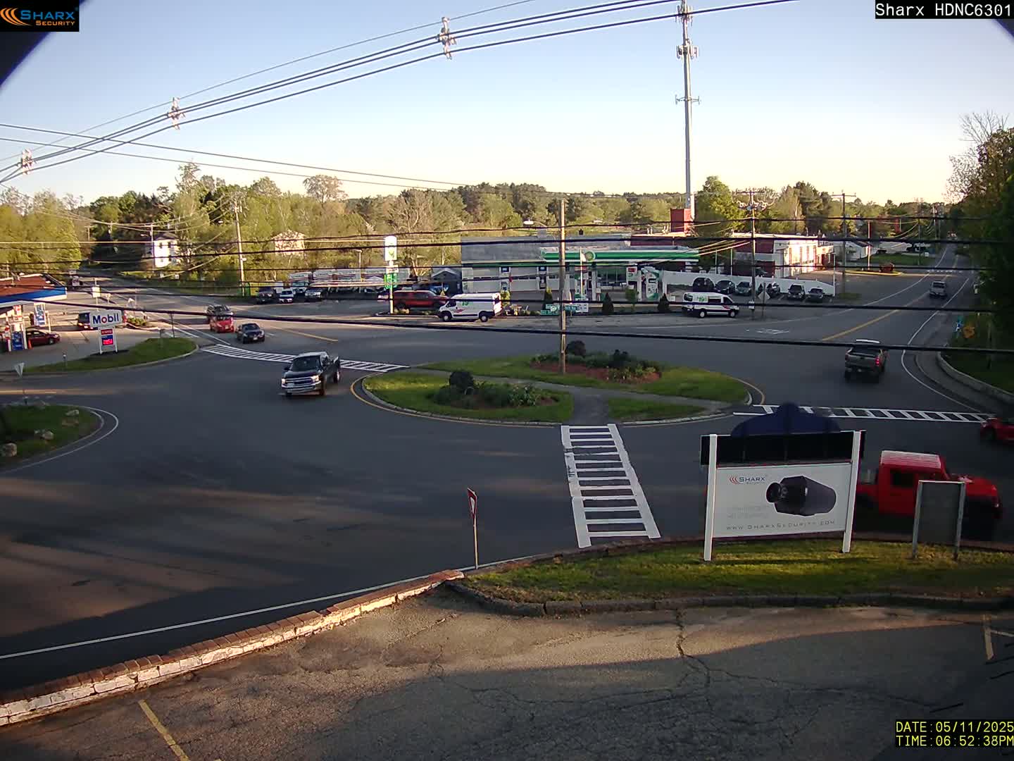 A sunny day shows a multi-lane roadway intersection with a traffic circle, several vehicles, buildings, and greenery.