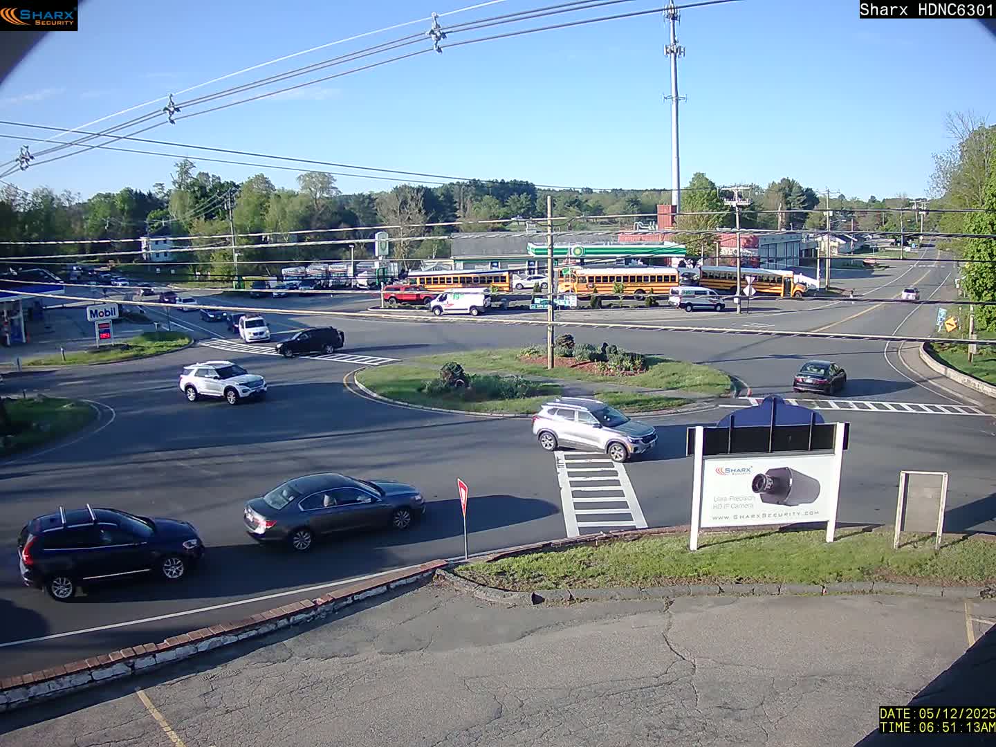 Several cars are driving around a roundabout near a gas station and school buses under a mostly sunny sky.
