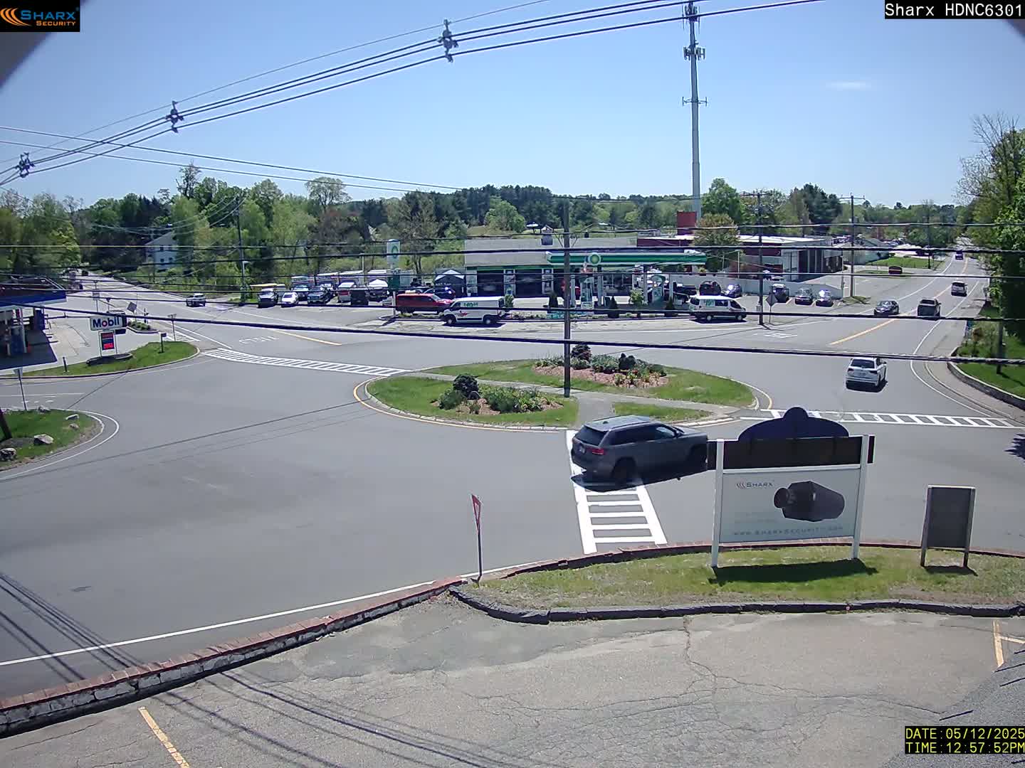 A sunny day reveals a traffic circle surrounded by a gas station, several parked cars, and a few vehicles driving through the intersection.