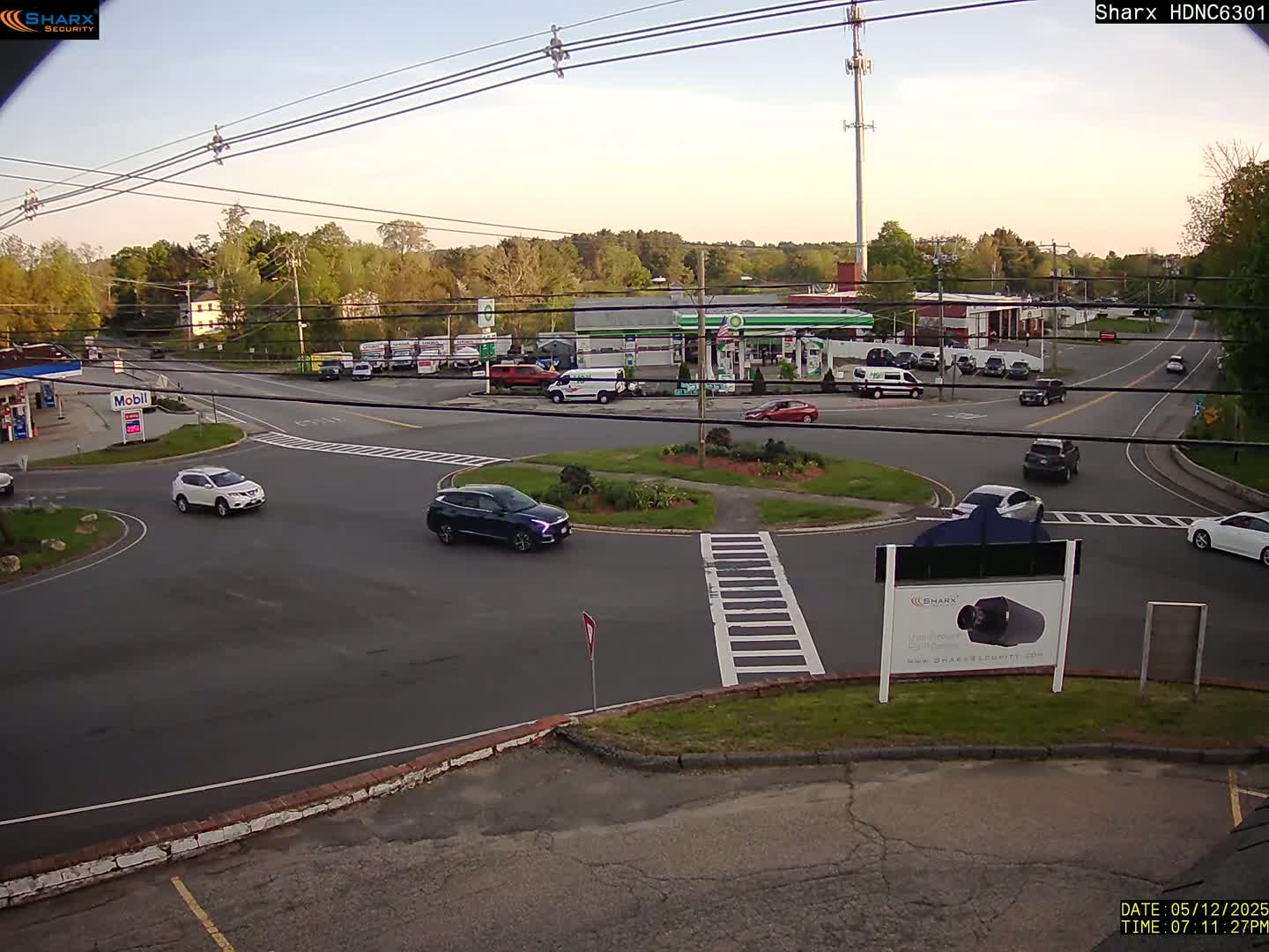A few cars are driving around a traffic circle near a gas station and several other buildings under a mostly clear sky.