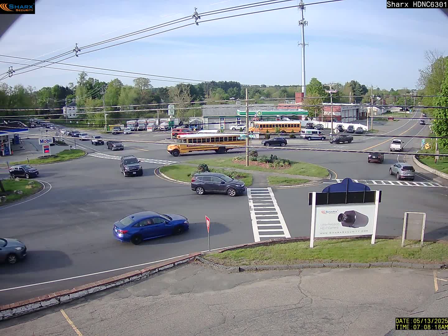 A sunny day shows a busy intersection with several cars and a school bus at a roundabout, surrounded by businesses and trees.