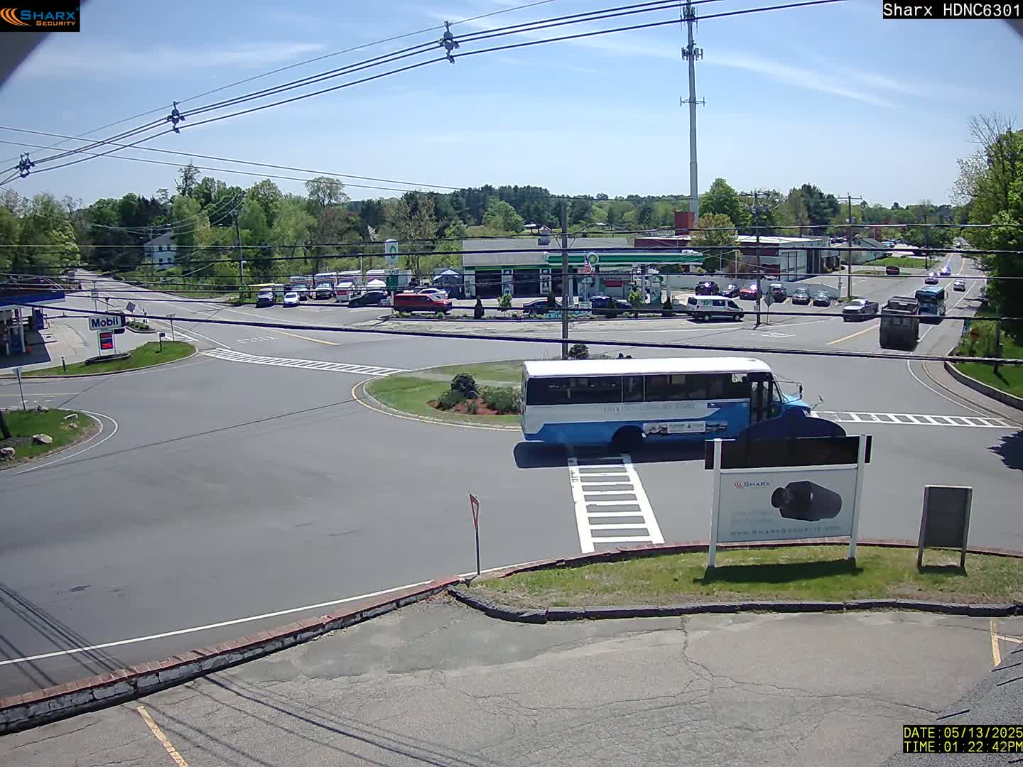 A blue and white bus is stopped at a multi-lane intersection on a sunny day, with several cars parked and driving nearby.