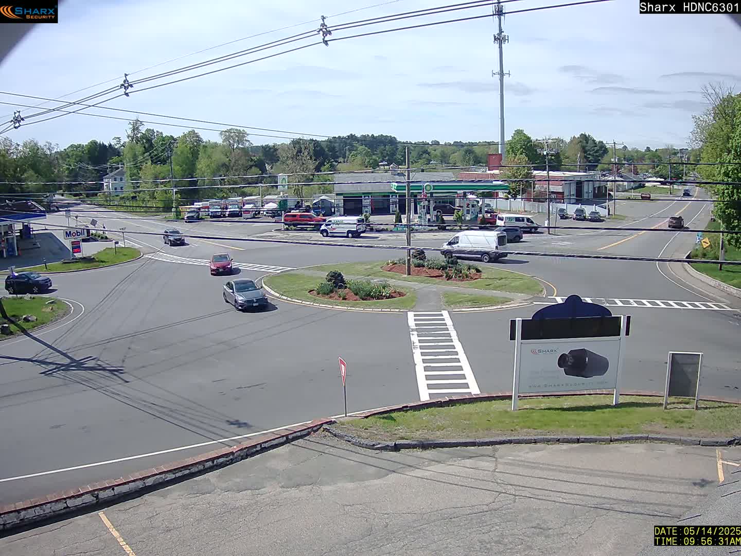 A sunny day shows several cars navigating a traffic circle near a gas station and other businesses, surrounded by trees and power lines.