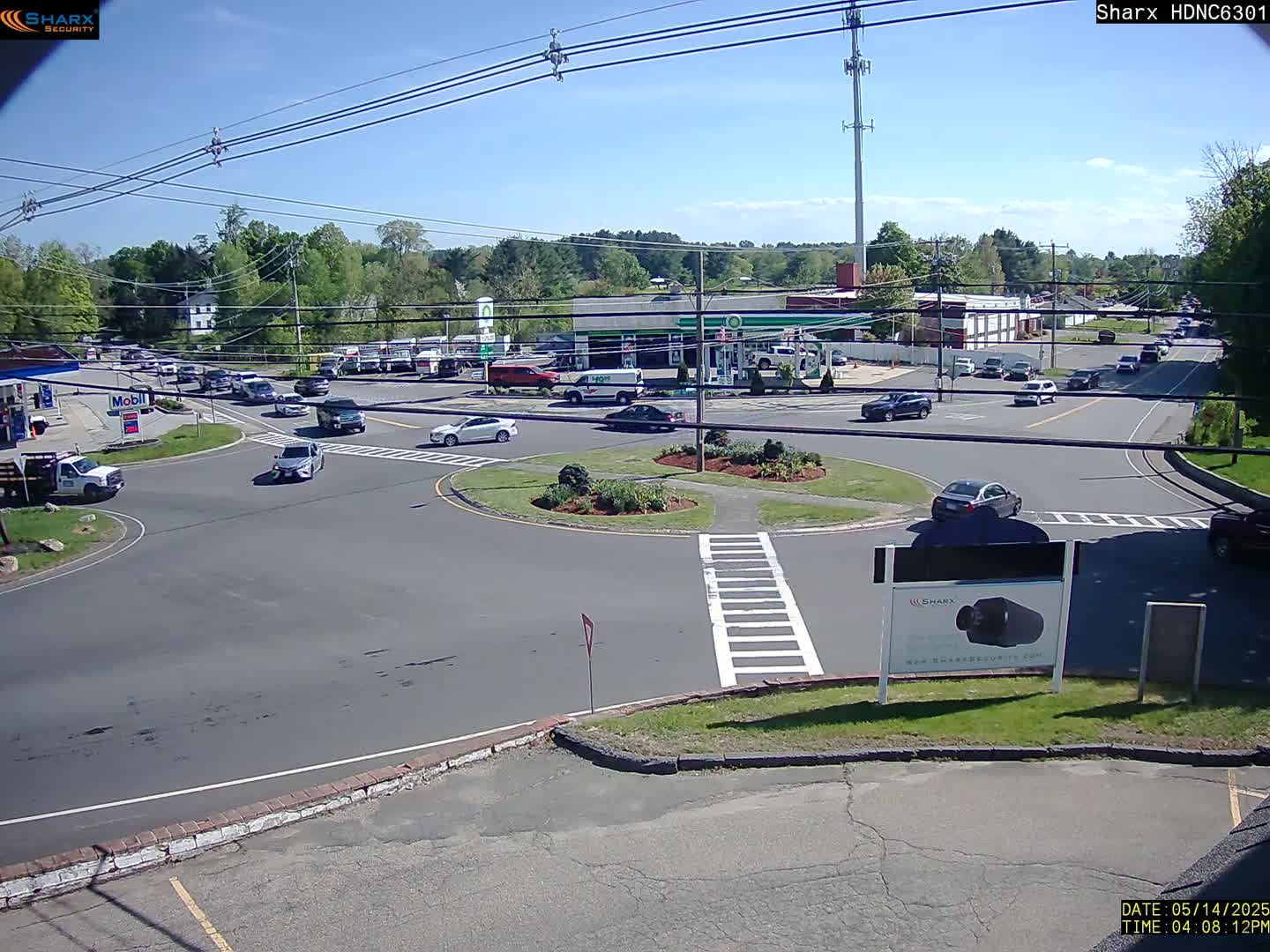 A sunny day shows several vehicles driving around a roundabout near a gas station and other buildings.