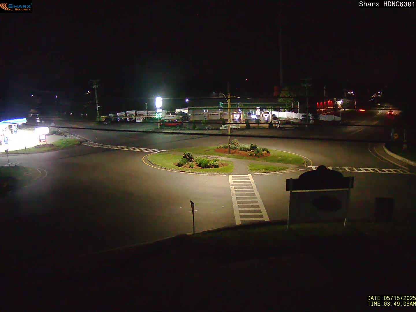 A nighttime view of a dark roadway intersection with a small traffic island, gas station, and other buildings in the background.
