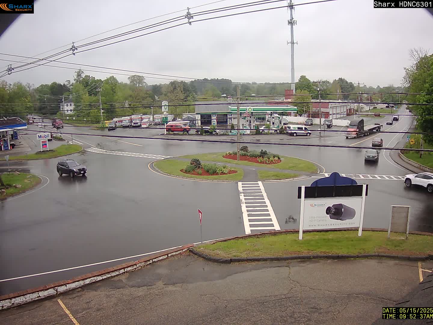 A wet roundabout in a town, surrounded by buildings, gas stations, and several vehicles, under an overcast sky.
