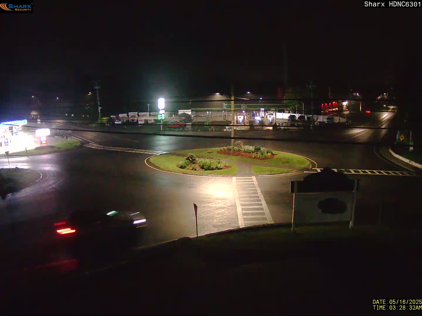 A wet, dark nighttime scene shows a roundabout with a few vehicles and buildings in the background, under artificial light.