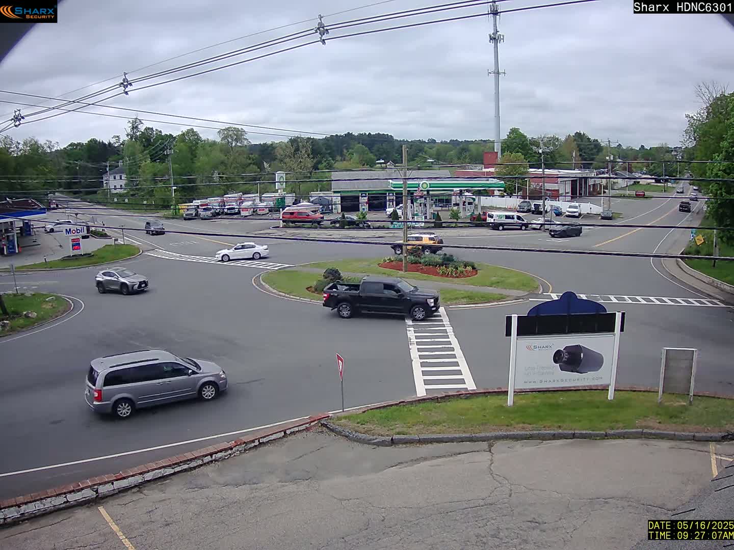 Several vehicles are driving around a traffic circle in front of a gas station and other businesses on an overcast day.