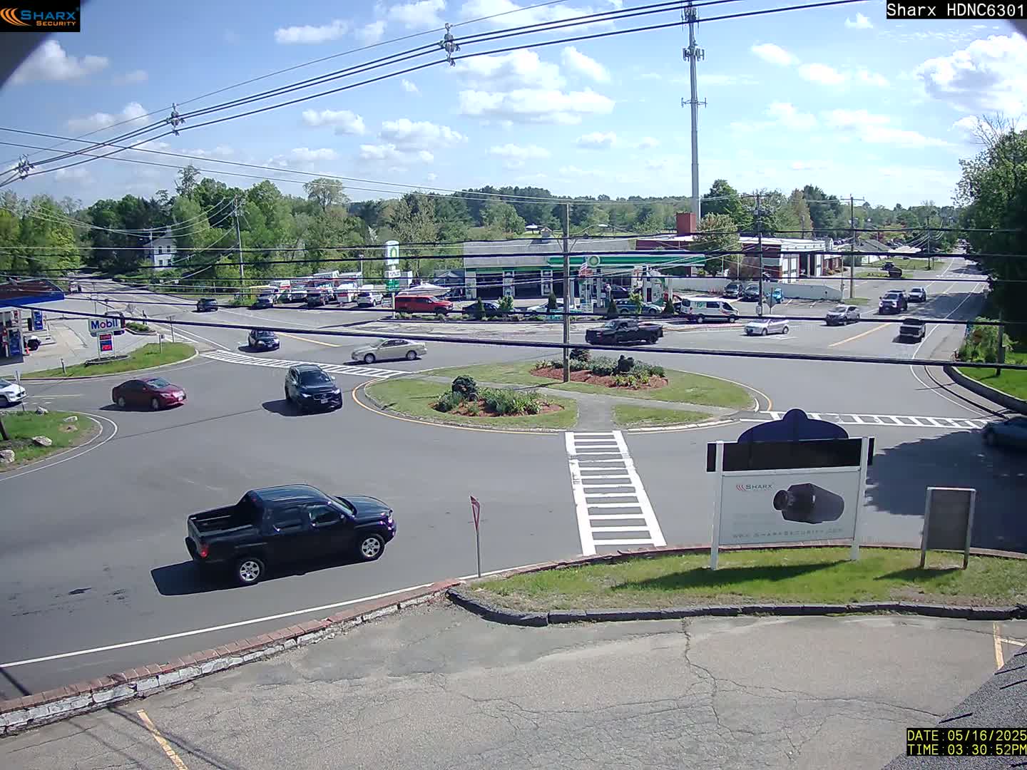 A sunny day shows a roundabout intersection with several cars and trucks, surrounded by businesses and trees.