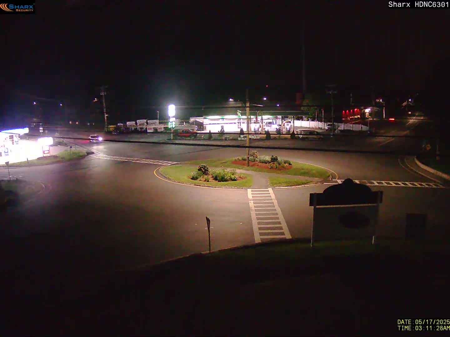 A nighttime view of a circular intersection with a gas station and other buildings in the background under dark, clear skies.