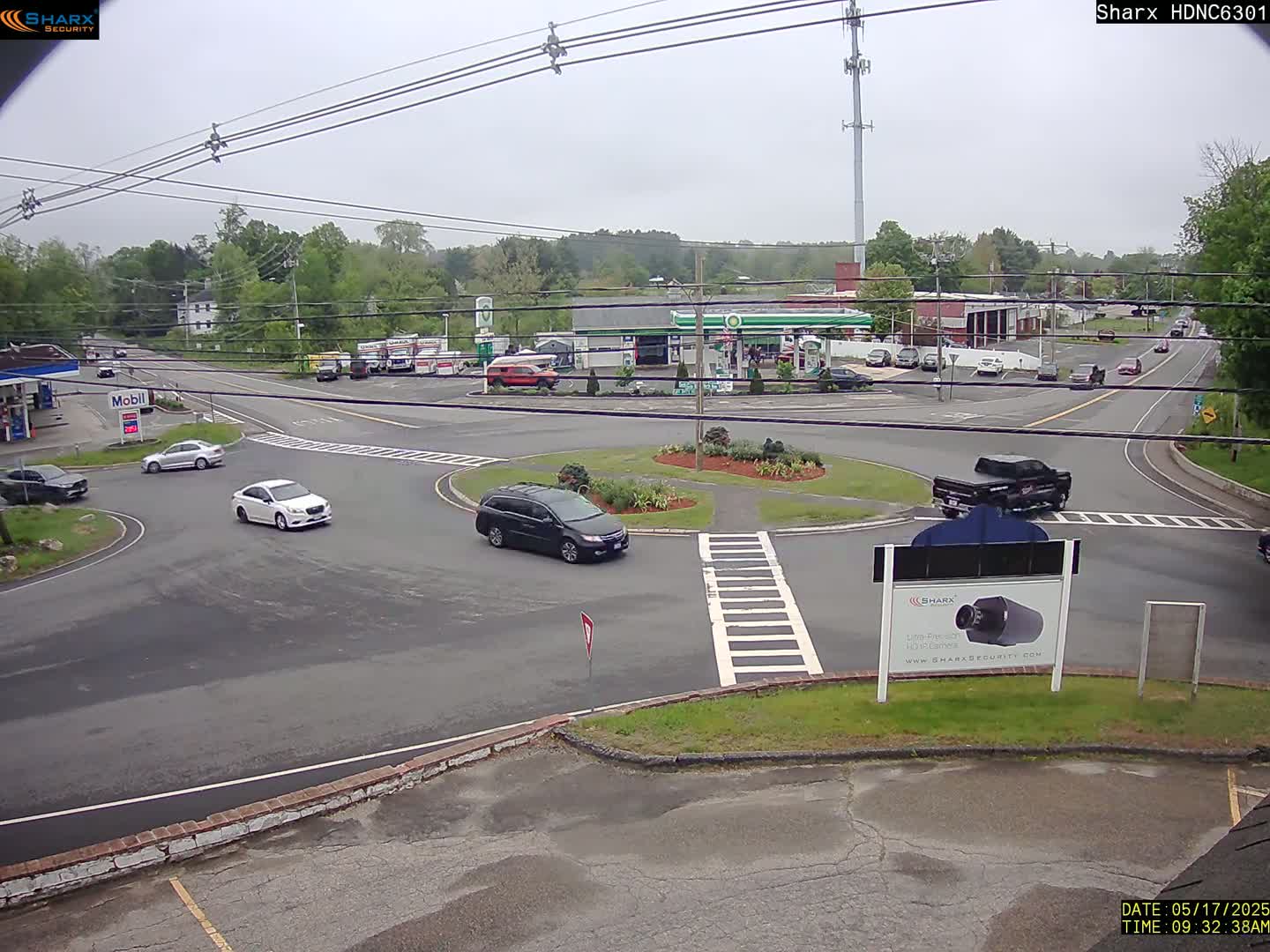 A slightly overcast day shows a traffic circle with several cars, and buildings including a gas station and other businesses, surrounded by green trees.