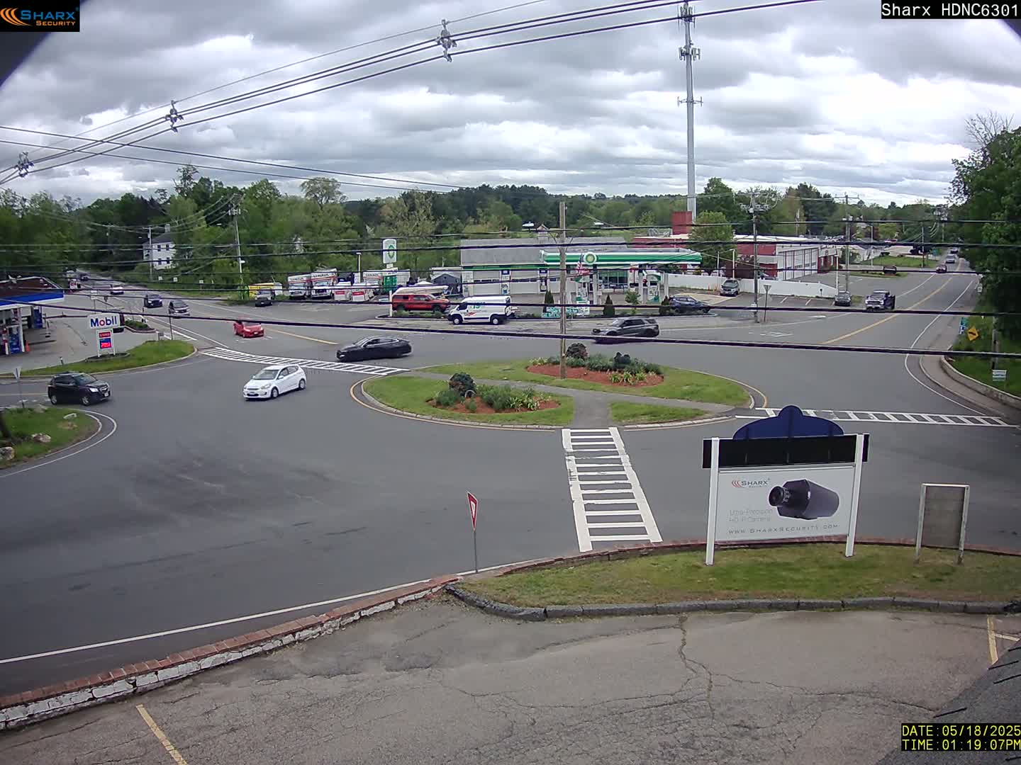 A mostly cloudy day shows a roundabout with several cars, a gas station, and other businesses, all surrounded by green trees.