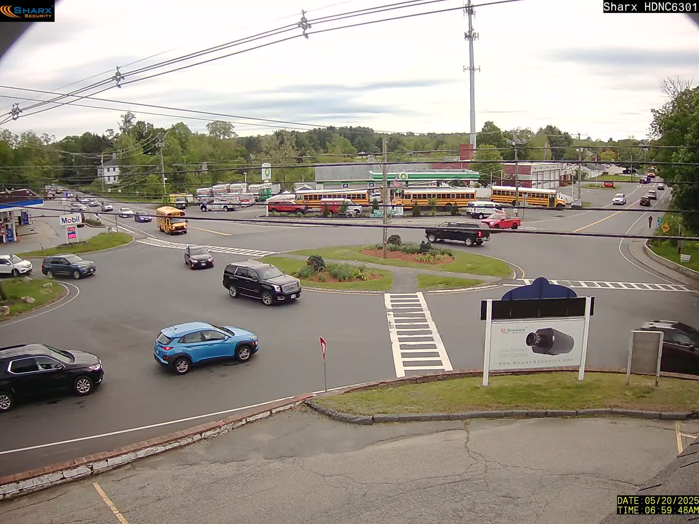 Several cars are navigating a roundabout near a gas station and several school buses under a partly cloudy sky.