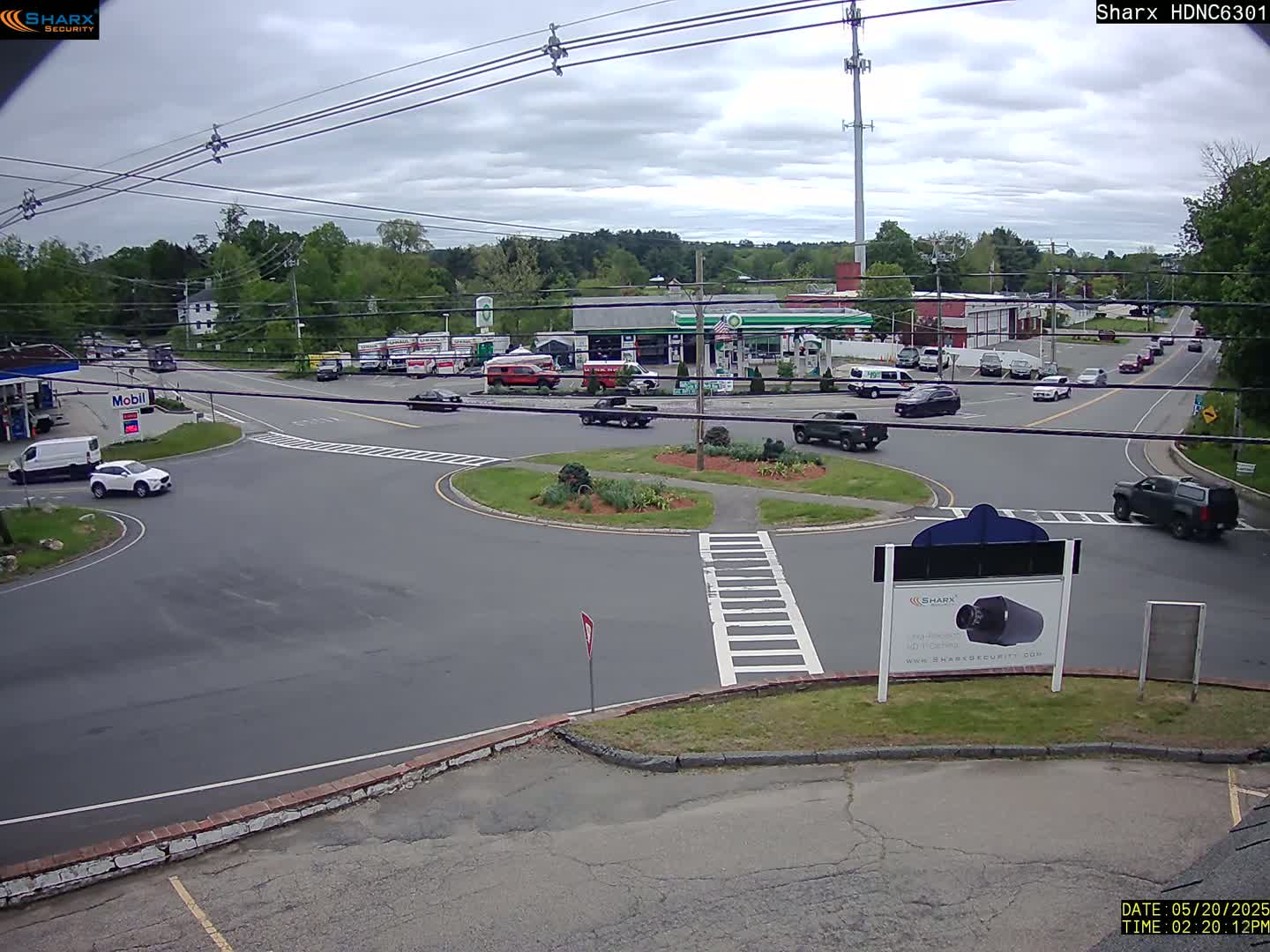 A mostly cloudy day reveals a roundabout intersection with several cars and trucks, surrounded by businesses and trees.